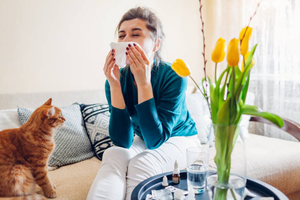 Woman sitting down and sneezing into a tissue with flowers and cat nearby